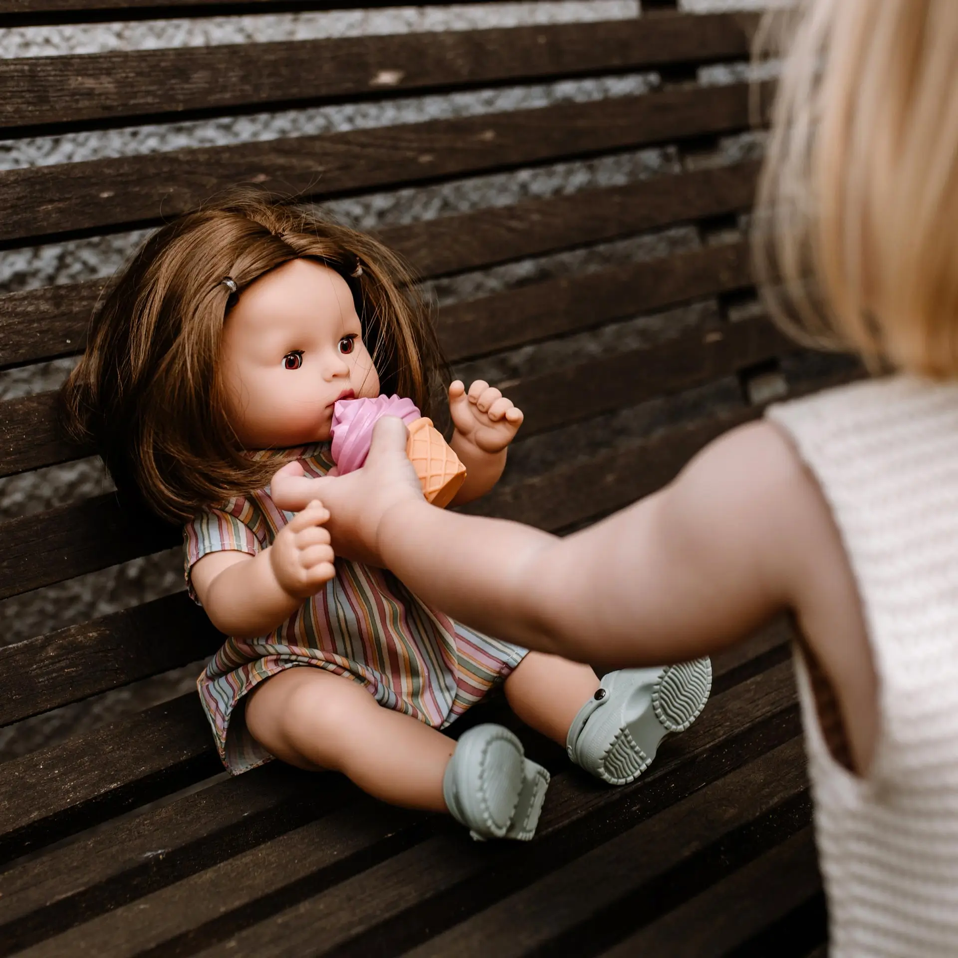 2518254 Maxy Aquini Beach Vibes Goetz Kind fuettert eine 42 cm große Babypuppe mit braunen Haaren und braunen Schlafaugen auf Sitzbank mit einem Eisspielzeug