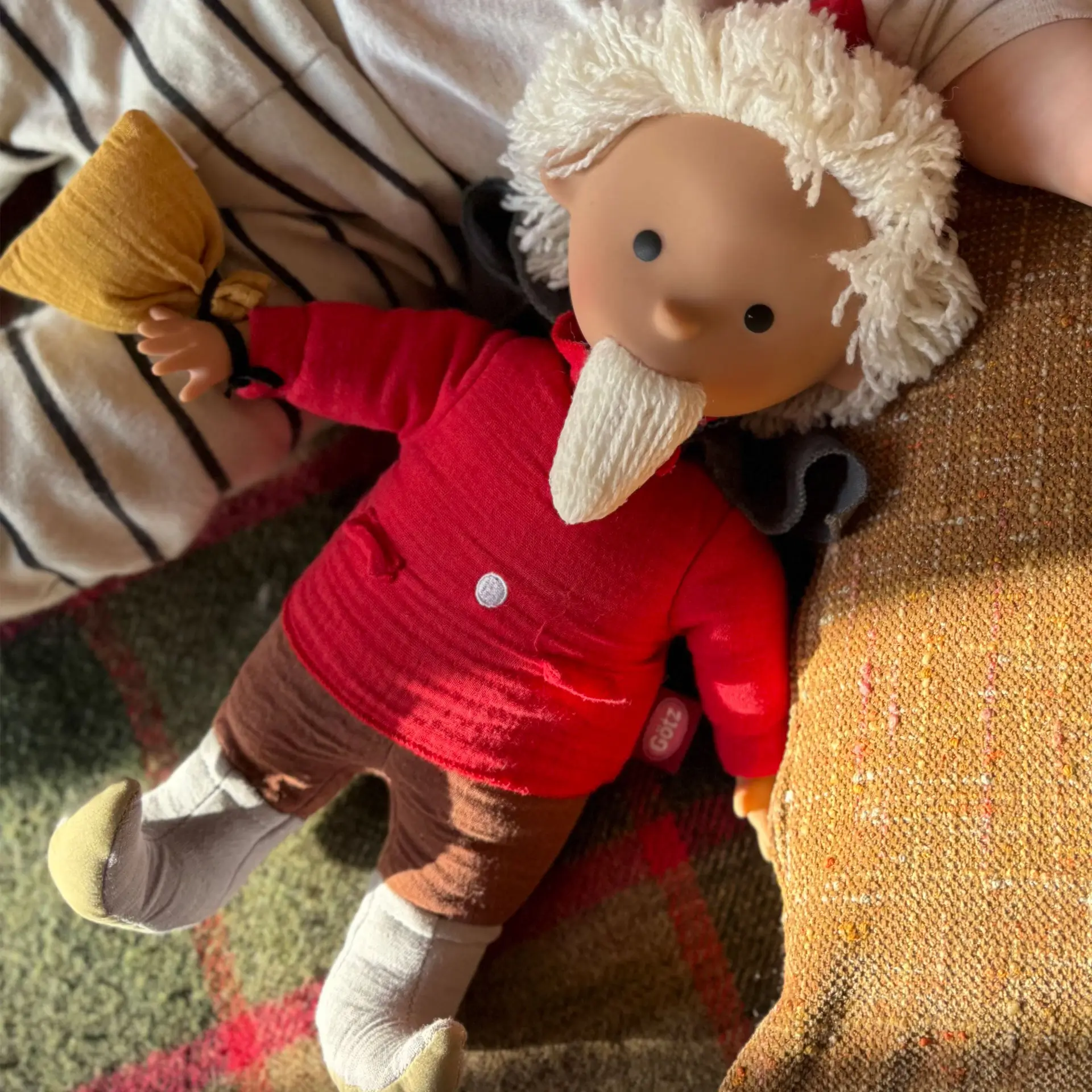 Sandmann Stoffpuppe mit rotem Pullover, brauner Hose, grauen Stiefeln und weißem Bart und Haaren liegt neben einem Kind