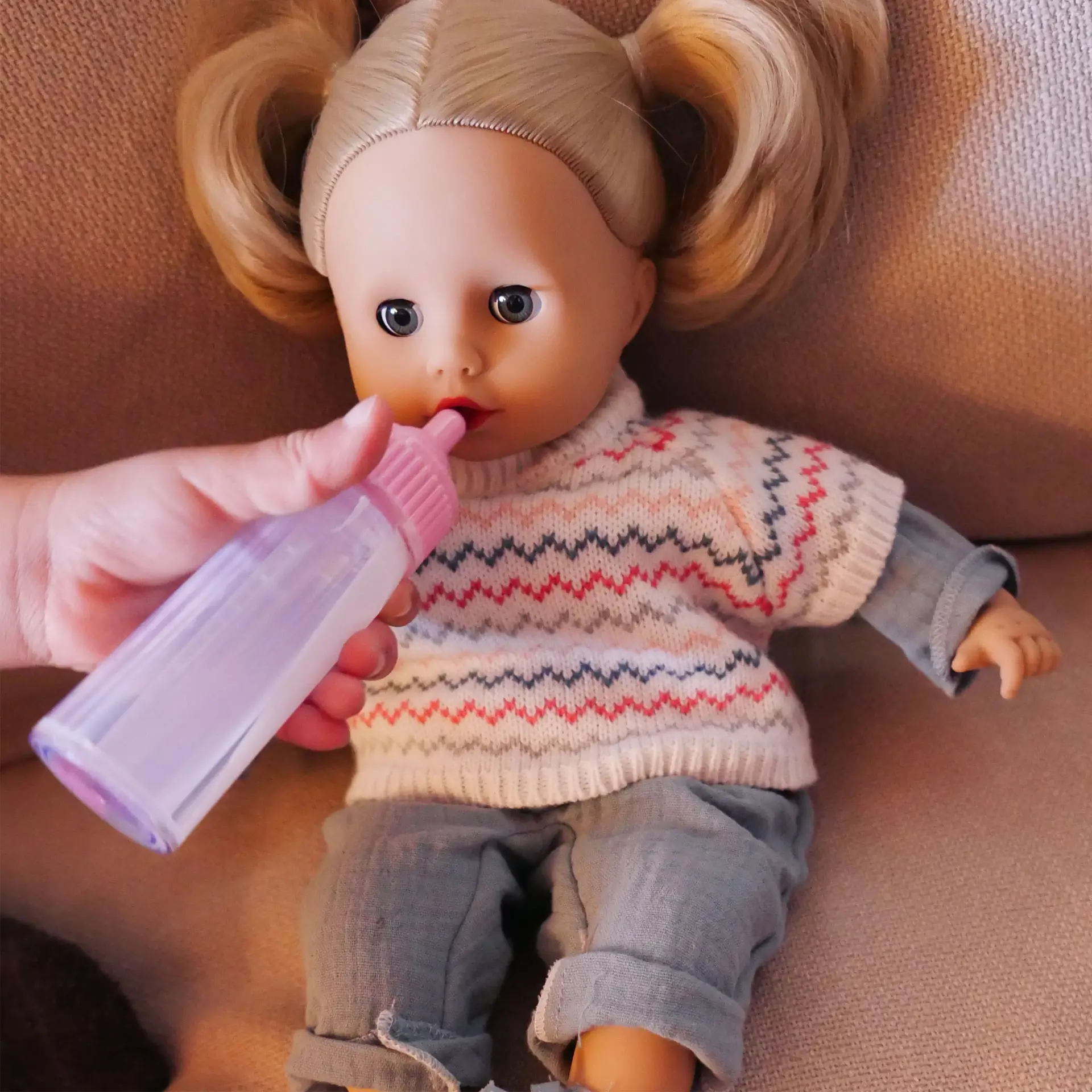 Kind fuettert 33 cm grosse Babypuppe mit blondem Haar und blauen Schlafaugen mit Milchflasche und traegt Overall mit Strickpullunder