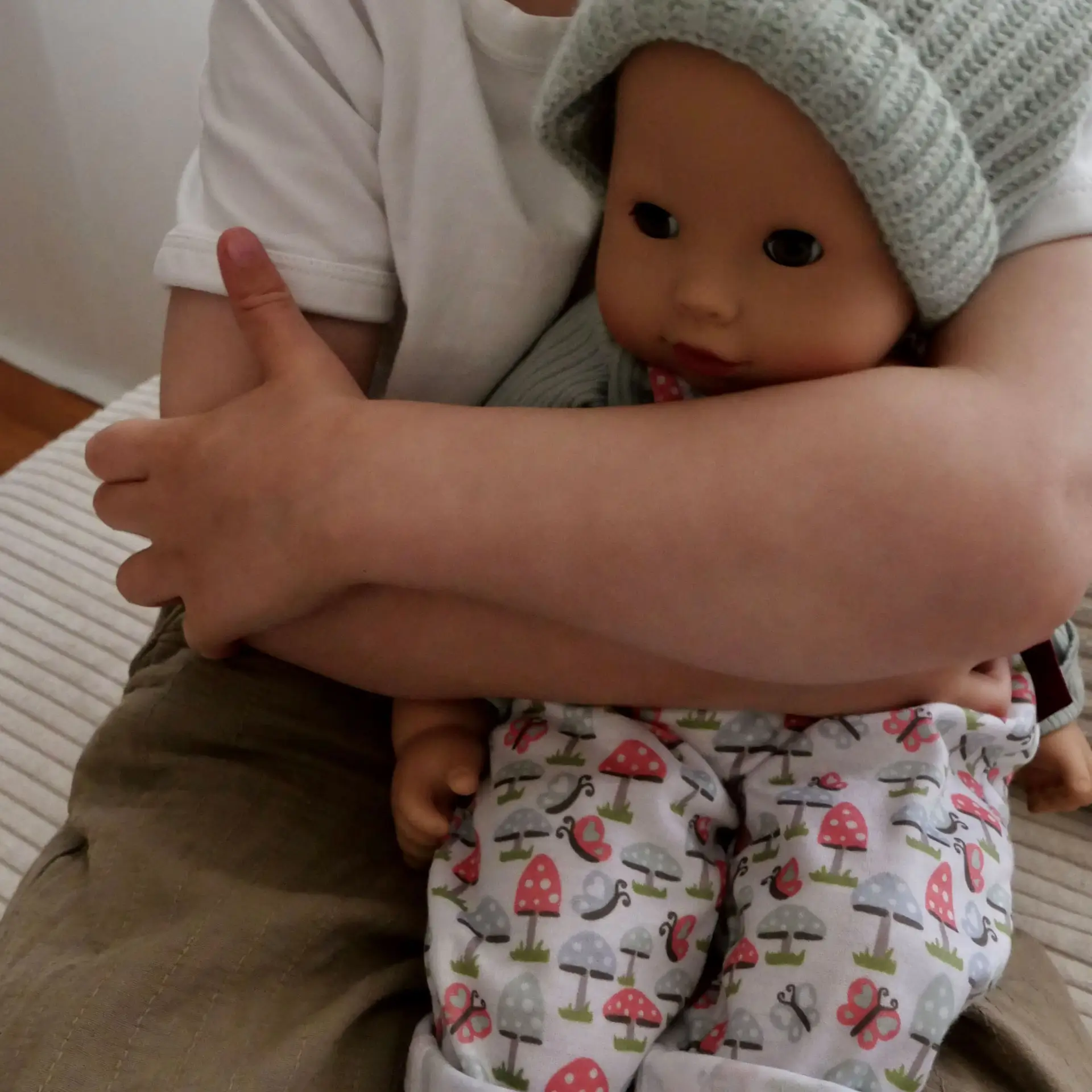  Kind haelt 42 cm grosse Babypuppe mit Schlafaugen im Arm, die eine Latzhose mit Pilzmustern traegt