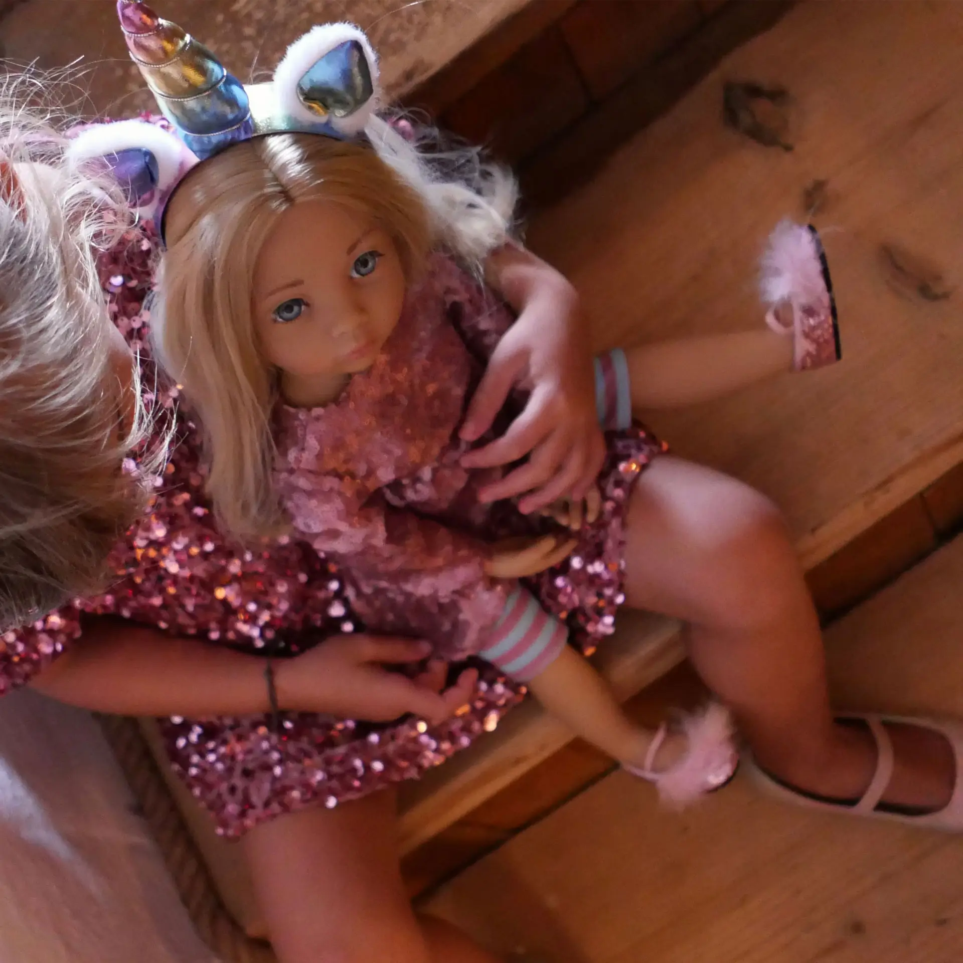 Kind haelt eine 50 cm grosse blonde Stehpuppe mit blauen Augen im Arm, die ein Einhorn-Outfit mit Pailletten, einen Einhorn-Haarreif und Ballerinas traegt