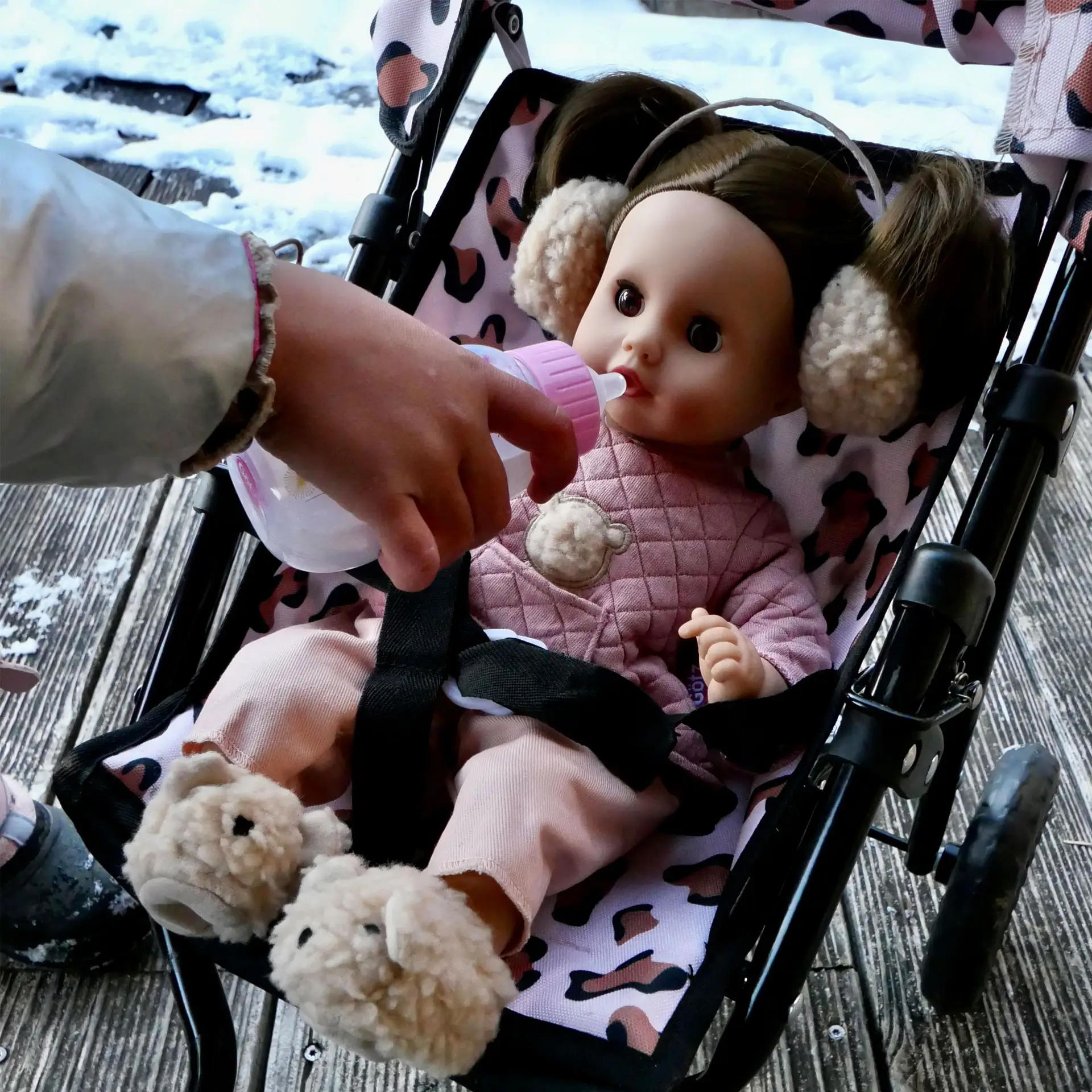 Eine 33 cm grosse Babypuppe mit braunen Haaren und braunen Schlafaugen sitzt im Buggy und trinkt aus einem Flaeschchen; sie traegt einen rosa Zweiteiler, Plueschohrenwaermer und Plueschhausschuhe