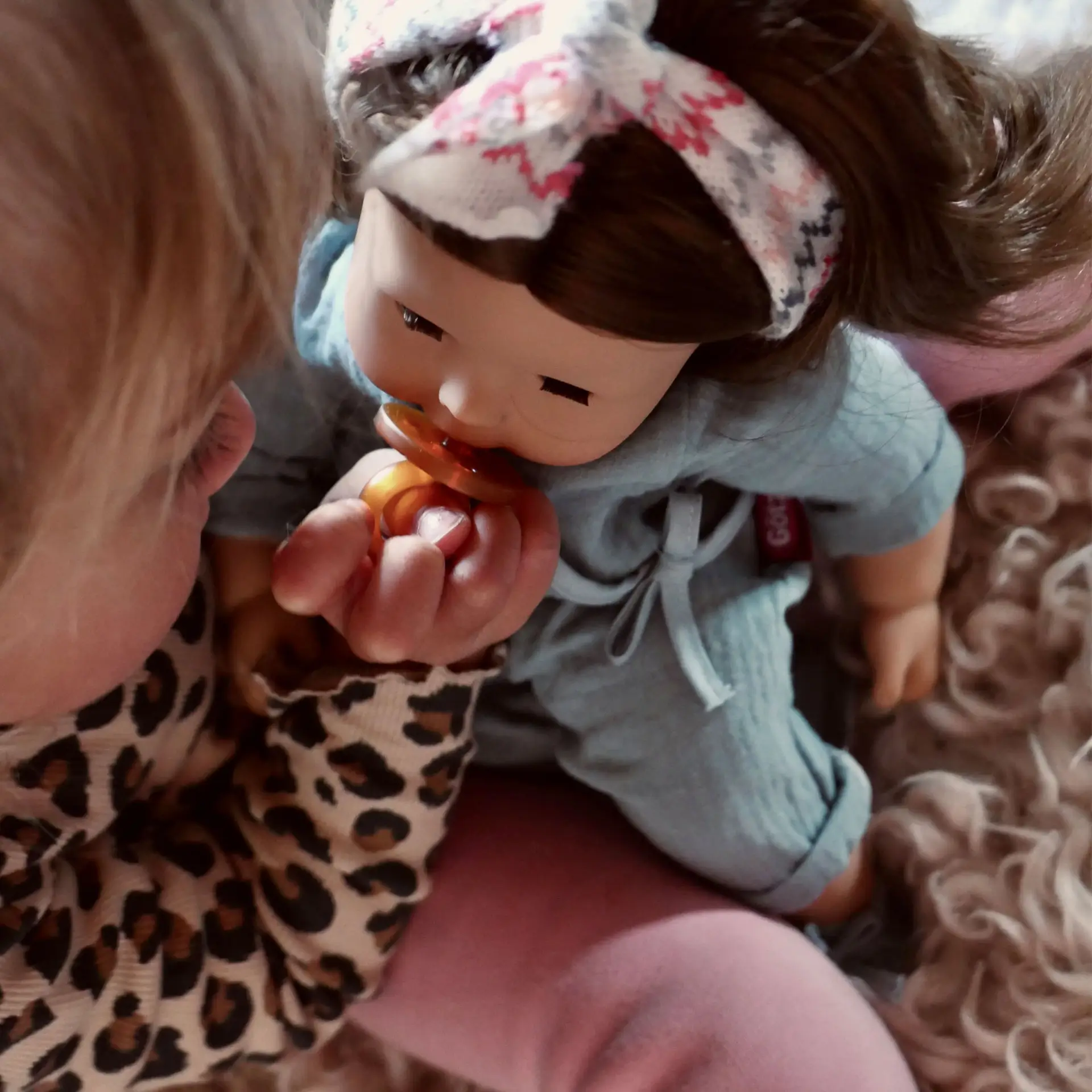 Kind gibt einer 42 cm grossen Babypuppe mit braunen Haaren und braunen Schlafaugen einen Schnuller in den Mund, welche einen blauen Anzug und ein Haarband traegt