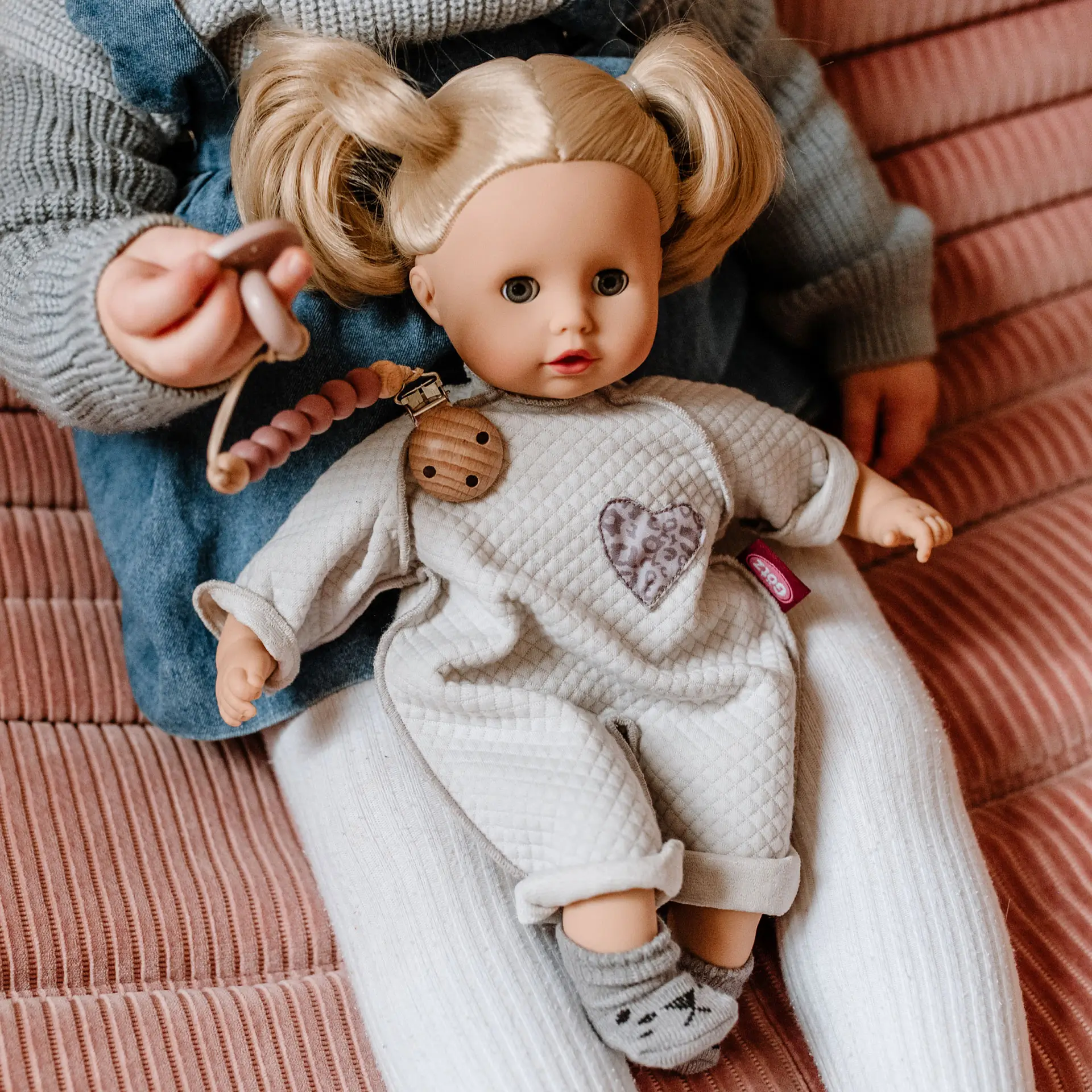 33 cm grosse blonde Babypuppe im grauen Overall sitzt auf dem Schoss eines Kindes