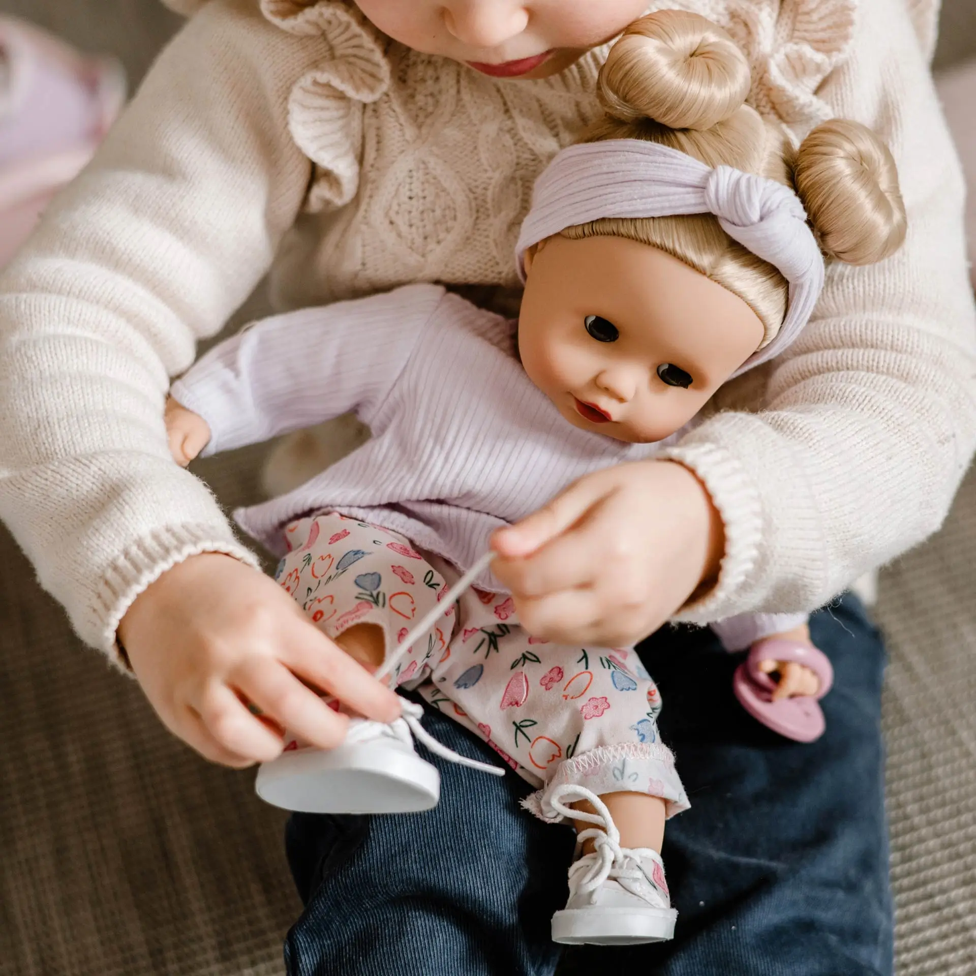 Kind bindet einer 33 cm grossen Babypuppe mit blauen Schlafaugen und blonden Haaren die Schuhe