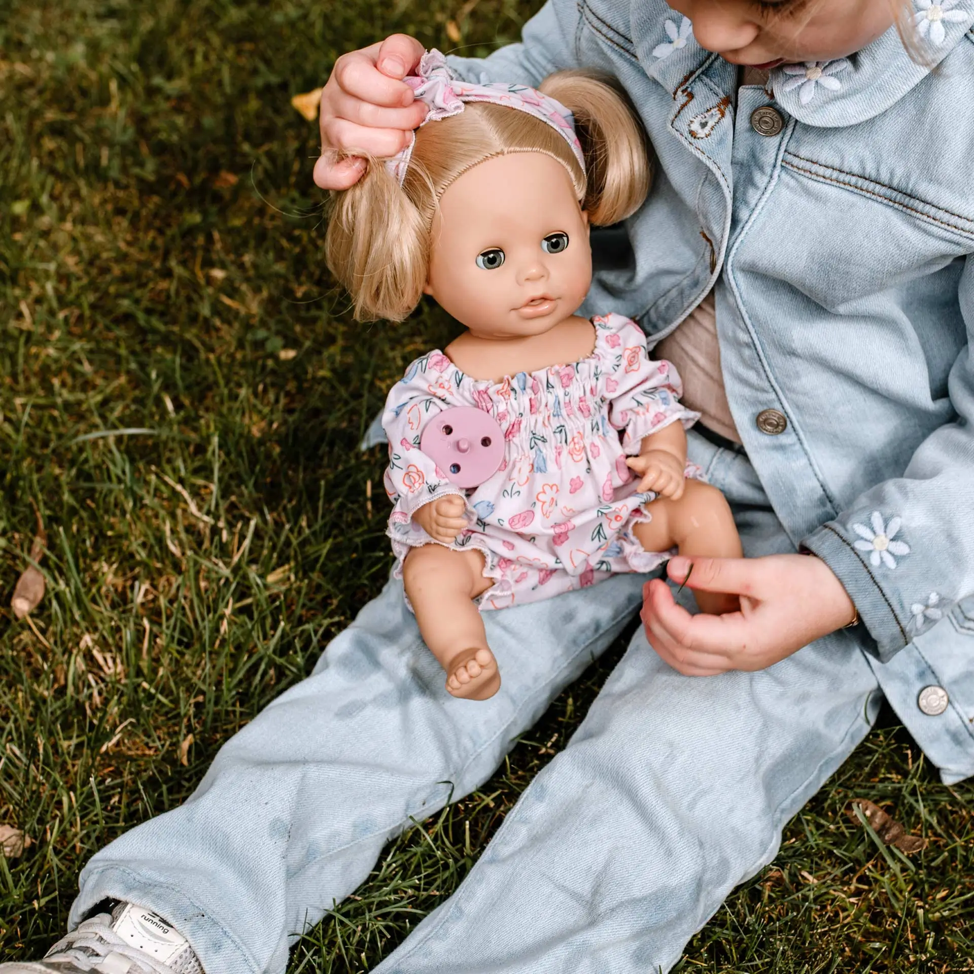 2353252 Sleepy Aquini Girl BliBlaBlume Goetz Kind sitzt auf der Wiese und haelt eine 33 cm große Babypuppe mit blonden Haaren Schlafaugen und Jumpsuit im Blumenprint