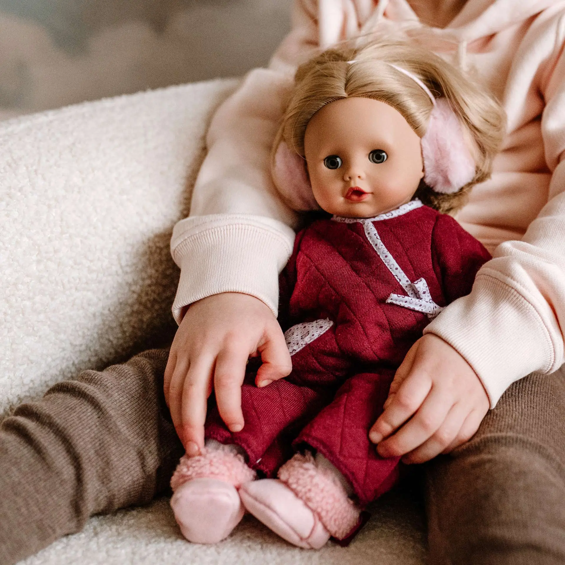 2520968  Muffin Jubilee Goetz Kind sitzt mit einer 33 cm großen Babypuppe mit blonden Haaren Schlafaugen und einem roten Overall auf dem Sofa