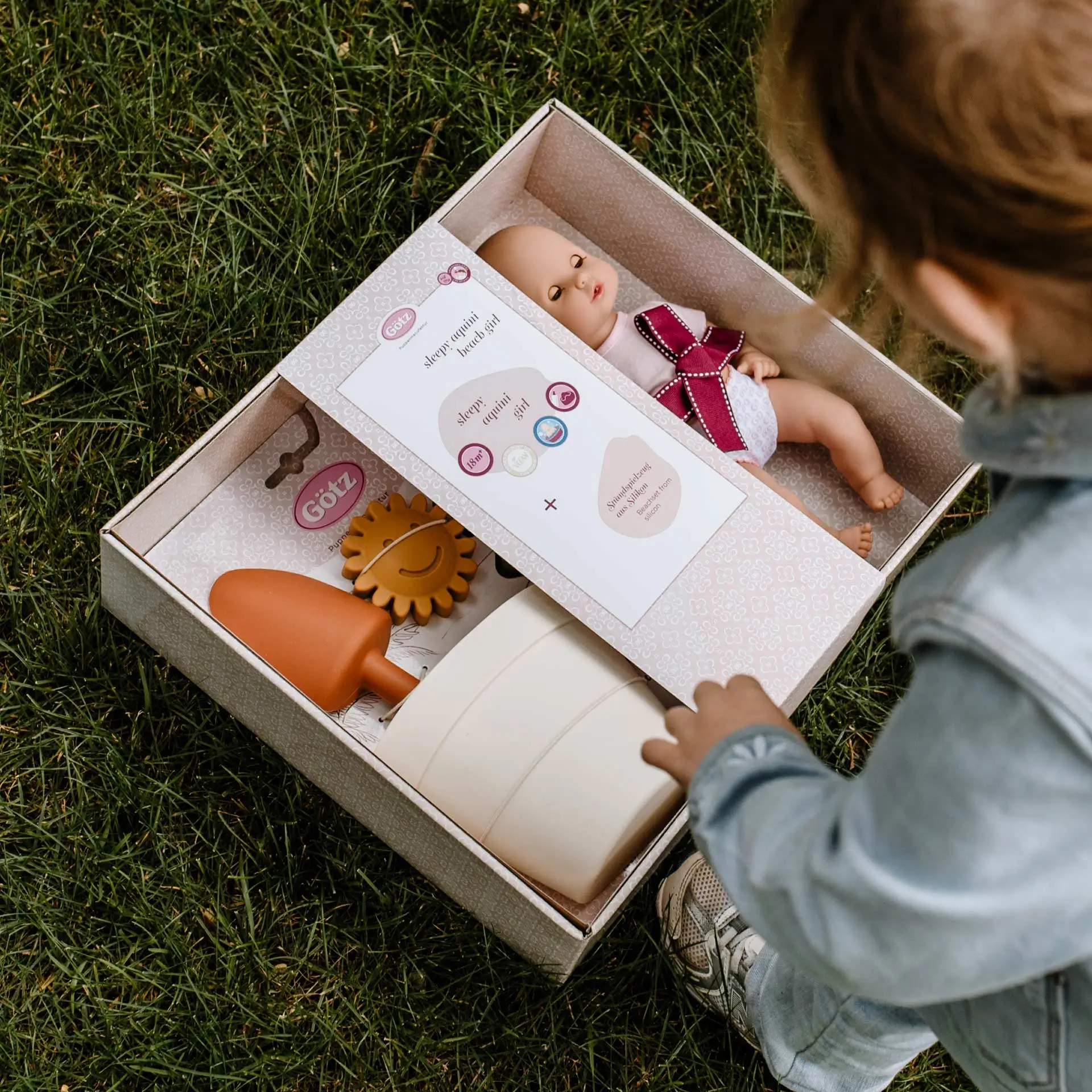 2453557 Geschenkbox Sleepy Aquini Beach Girl Goetz Kind oeffnet eine Geschenkbox, in der eine 33 cm grosse Badepuppe mit Schlafaugen und Sandspielzeug liegt
