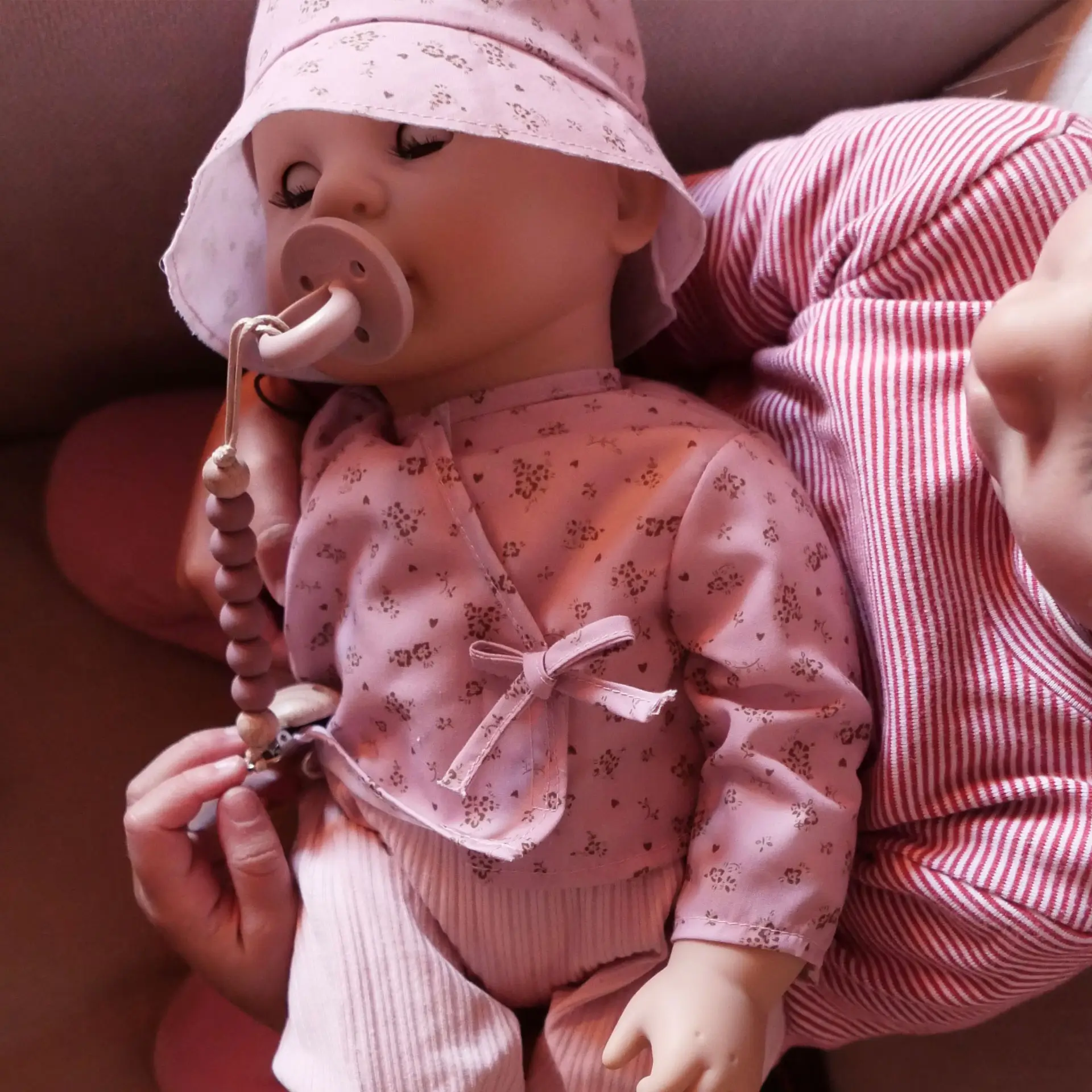 Kind haelt eine 48 cm grosse Babypuppe mit braunen Schlafaugen im Arm. Die Puppe traegt einen rosa Anzug mit Blumenmuster, ein Cappy sowie einen Schnuller mit Schnullerkette