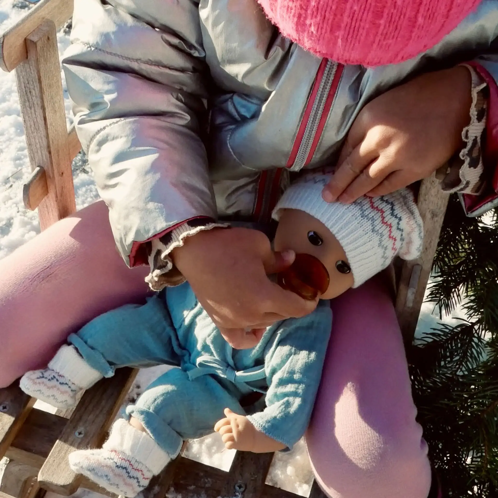 Kind sitzt auf einem Schlitten und hält eine 33 cm grosse Babypuppe mit blauen Schlafaugen, die einen blauen Anzug mit Strickschuhen und Strickmuetze traegt