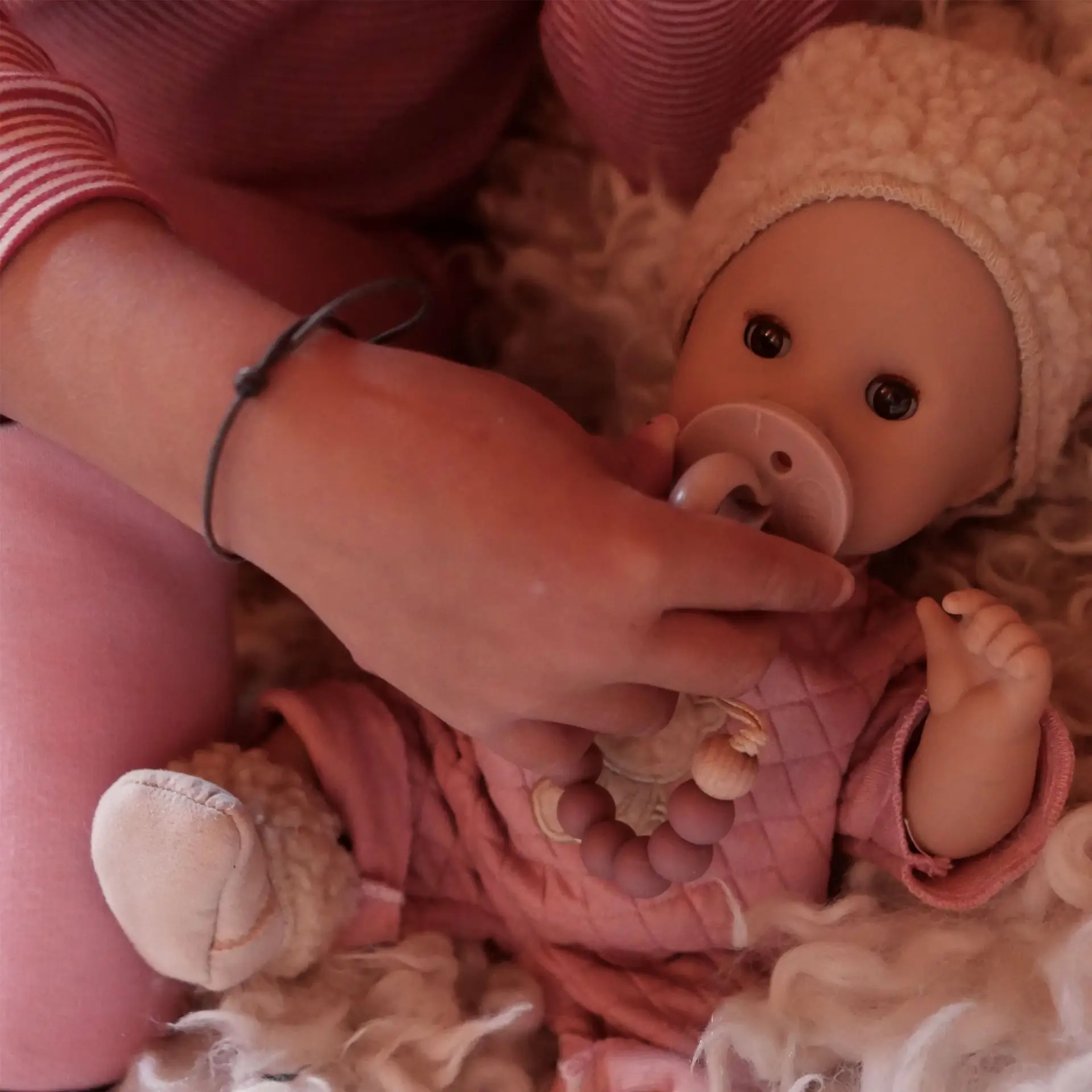 Kind gibt einer 33 cm grossen Badepuppe mit braunen Schlafaugen einen Schnuller in den Mund; die Puppe traegt einen rosa Anzug, eine Plueschmuetze und Plueschboots