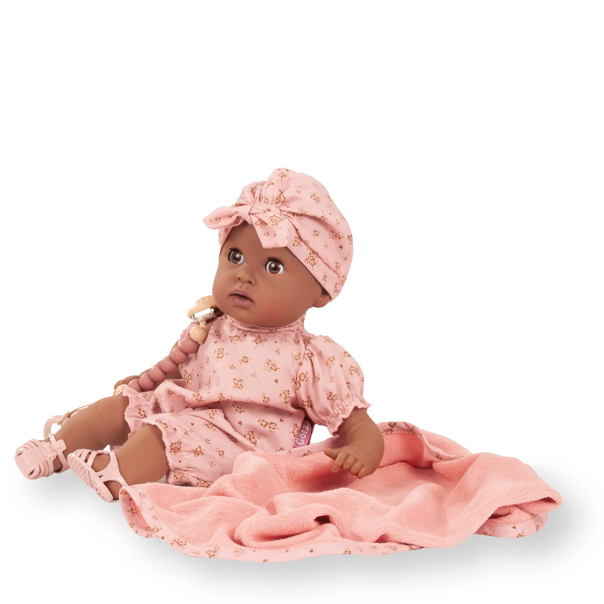 Badepuppe Gr. 33 cm in braunem Vinyl, ohne Haare mit braunen Schlafaugen. Sie traegt einen rosafarbenen Sommerstrampler mit goldenen Bluemchen und einer Muetze. Dazu ein Schnuller mit Schnullerkette, dazu ein Kapuzentuch und Badesandalen von Goetz