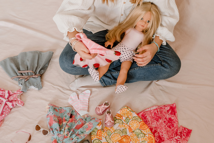 Kind sitzt mit 46 cm großer Stehpuppe mit Weichkörper und blonden Haaren auf dem Schoß umgeben von Puppenkleidern