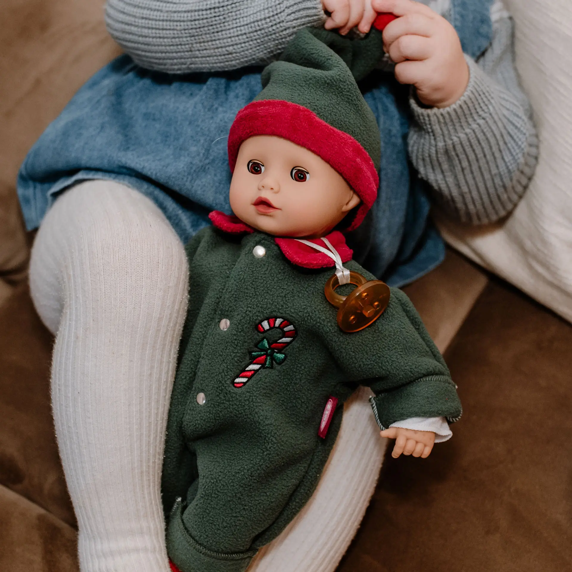 Kind spielt mit 33 cm grosser Babypuppe mit Schlafaugen, die einen festlichen gruen-roten Fleeceanzug und eine Zipfelmuetze traegt