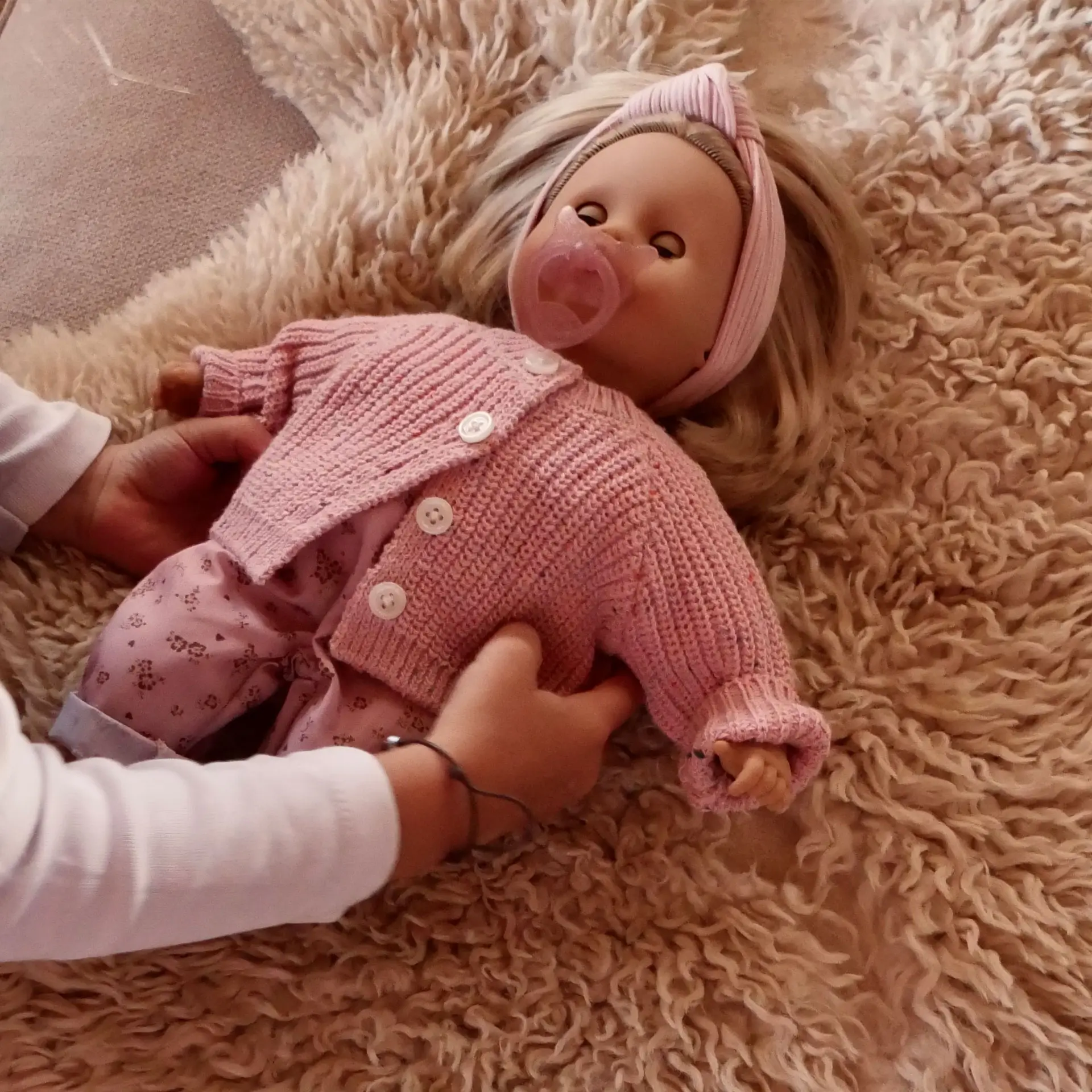 Kind legt eine 33 cm grosse Babypuppe mit blonden Haaren und blauen Schlafaugen auf den Teppich; die Puppe traegt eine rosa Strickjacke, ein Haarband und hat einen Schnuller im Mund