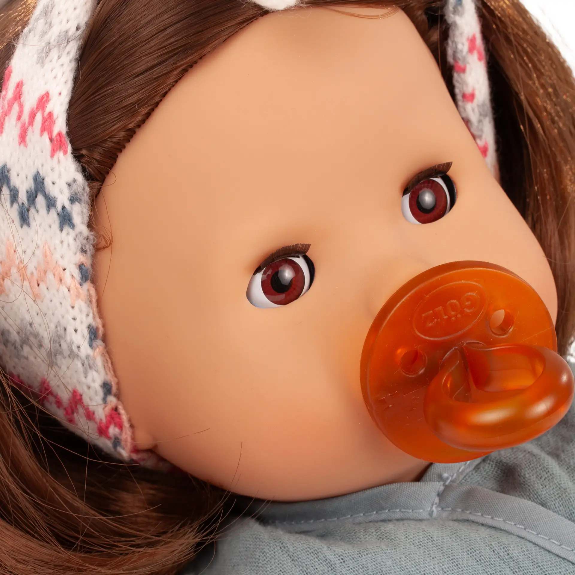 Portrait von Babypuppe Gr. 42 cm mit braunen Augen und braunem Haar. Sie trägt das Strickhaarband im Zick-zack-Muster und einem Schnuller von Goetz