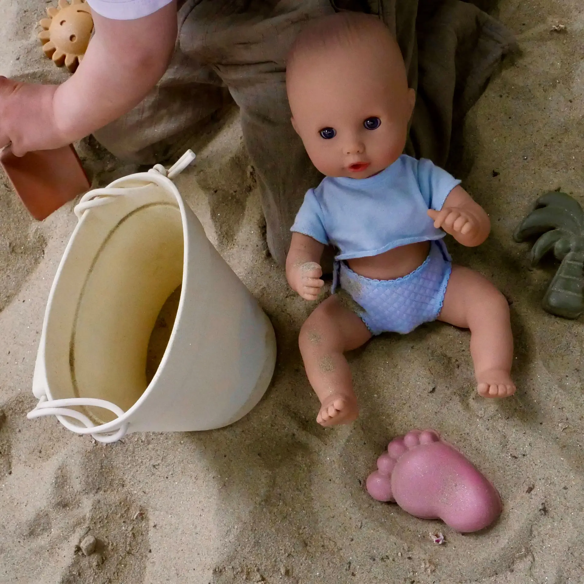 Kind spielt im Sand mit 33 cm grosser Badepuppe mit blauem Unterhemd Stoffwindel und Sandspielzeug