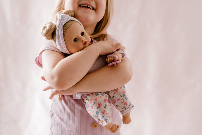 Maedchen umarmt eine 33 cm große Babypuppe mit blonden Haaren und laechelt