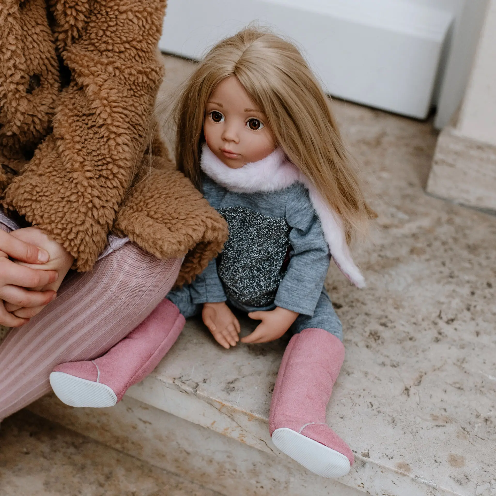 Kind sitzt neben 50 cm grosser blonder Stehpuppe mit rosa Stiefeln, grauer Hose und grauem Oberteil