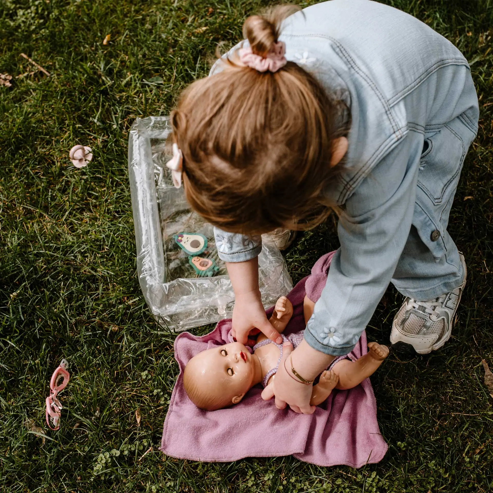 Kind legt eine 33 cm große Babypuppe mit Schlafaugen auf ein Kapuzentuch auf die Wiese 