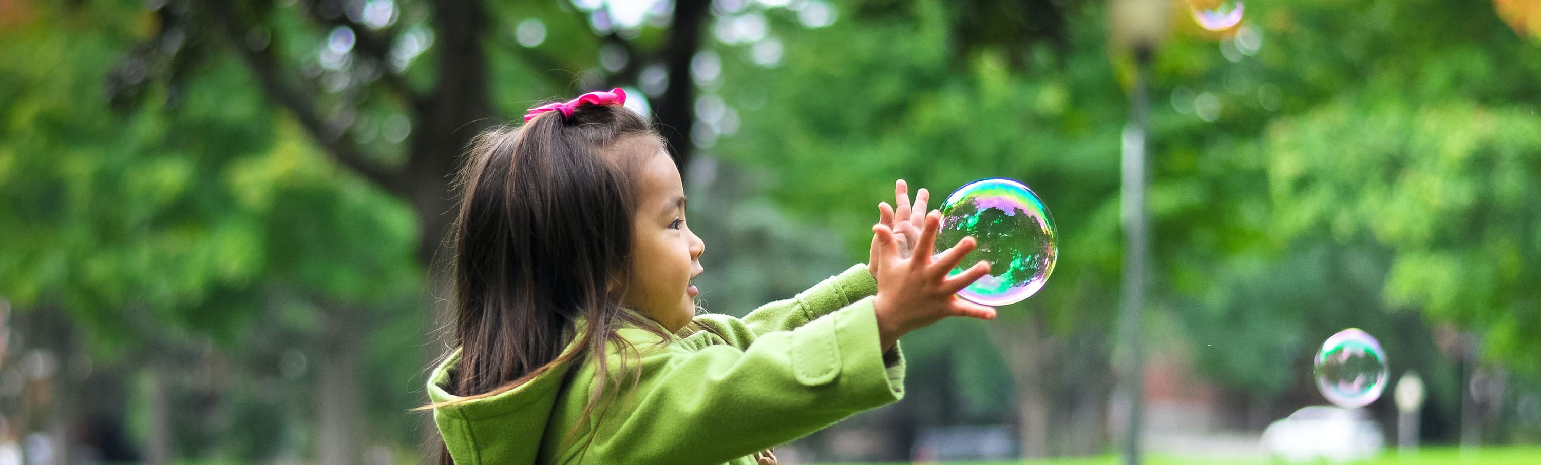 Kind mit gruener Jacke und braunen Haaren streckt die Hand nach einer Seifenblase aus