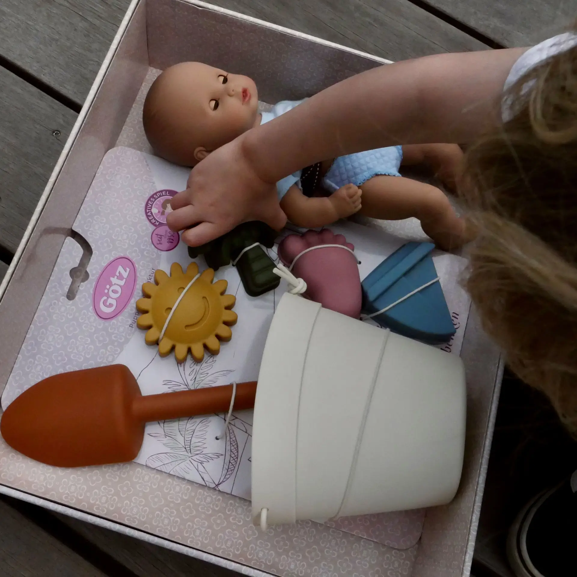 Kind oeffnet eine Geschenkbox, in der eine 33 cm grosse Badepuppe mit Schlafaugen und Sandspielzeug liegt