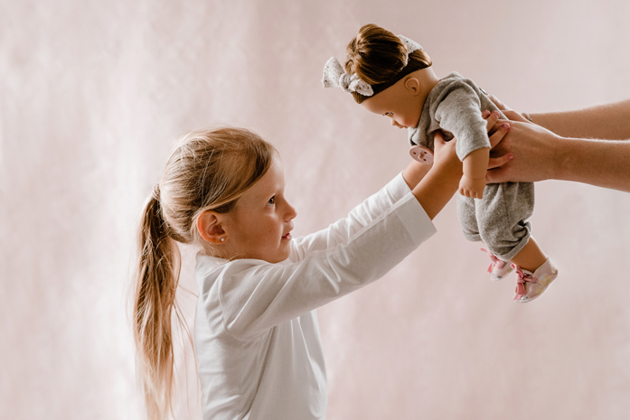 Kind haelt eine 42 cm grosse Babypuppe mit braunen Haaren im grauen Overall in die Luft